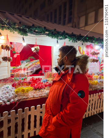 Handsome millennial man model with a hairstyle in winter red jacket walks in night city with festive lights and Christmas market. Christmas and winter holidays season Handsome millennial man model with a hairstyle in winter red jacket walks in night city with festive lights and Christmas market. Christmas and winter holidays season 118790451