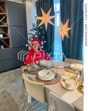 Millennial woman in a Santa suit sits thoughtfully and sad at a festive dining table, surrounded by Christmas decorations and a lit tree. Moment of holiday reflection and problem loneliness in Millennial woman in a Santa suit sits thoughtfully and sad at a festive dining table, surrounded by Christmas decorations and a lit tree. Moment of holiday reflection and problem loneliness in 118790470