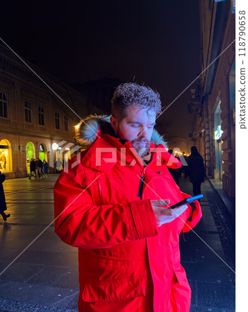 Millennial man in vivid red parka stands on city street at night, engrossed in their smartphone. Illuminated by the warm glow of city lights, capturing essence of urban life and digital connectivity 118790658