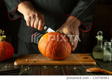 A chef cuts pumpkin with a knife on a wooden board in a restaurant kitchen. The process of preparing ingredients for a seasonal fall dish or pie. American national dish 118790961