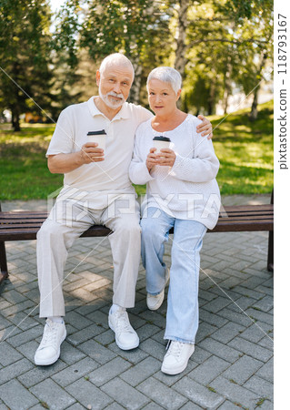 Full length vertical portrait of loving senior couple enjoying coffee sitting hugging on park bench, basking in summer warmth, exuding love and happiness, embodying love and happiness in retirement. Full length vertical portrait of loving senior couple enjoying coffee sitting hugging on park bench, basking in summer warmth, exuding love and happiness, embodying love and happiness in retirement. 118793167
