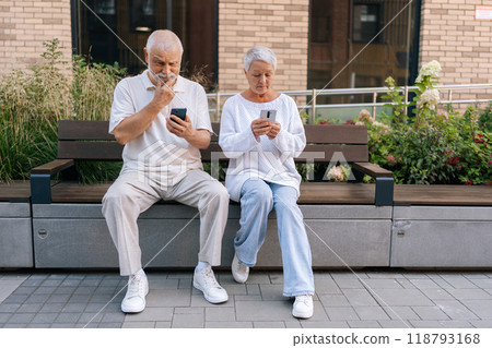 Portrait of elderly married couple enjoying sunny summer day browsing smartphones sitting on urban bench. Gray-haired senior man and woman using mobile phone smiling looking to screen. 118793168