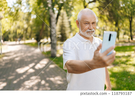 Portrait of handsome white-bearded senior man taking selfies on smartphone while walking on summer sunny park surrounded by lush greenery and trees. Happy elderly male capturing selfie in park. 118793187