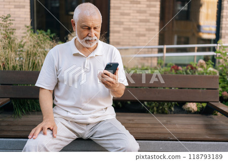 Portrait of handsome senior man with white beard using smartphone focused looking at screen sitting on urban bench on summer day. Relaxed gray-haired elderly grandfather using mobile phone outdoors. Portrait of handsome senior man with white beard using smartphone focused looking at screen sitting on urban bench on summer day. Relaxed gray-haired elderly grandfather using mobile phone outdoors. 118793189