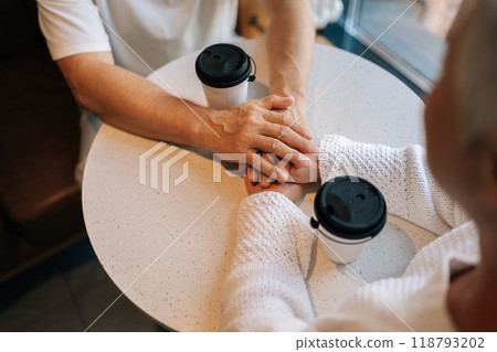 Closeup top view of unrecognizable happy senior couple sharing warm conversation over coffee, holding hands intertwined, radiating love and companionship in cozy cafe. Concept of active aging. 118793202
