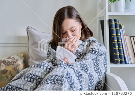 Young sick woman sitting under warm blanket sneezing coughing in handkerchief 118793270