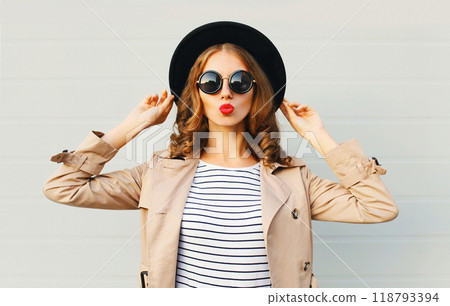 Fashion portrait young woman blowing kiss wearing a black hat sunglasses coat over grey background 118793394
