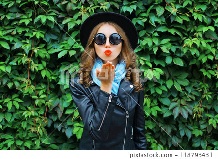 Fashion portrait young woman blowing a kiss in black hat over green leaves background 118793431