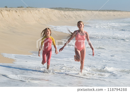Two girls are running joyfully along the shoreline, splashing through the shallow waves on a sunny beach. The sandy dunes and clear sky create a perfect summer day atmosphere full of fun. 118794288