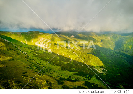 Breathtaking aerial view of green, rugged mountain peaks under dramatic sky. Scene captures contrast between vibrant landscape and moody clouds, with sunlight breaking through. Carpathians, Chornogora 118801545