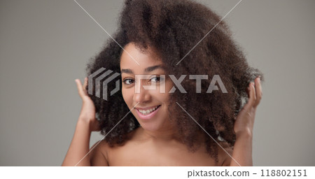 Beautiful African American woman with curly afro hairstyle and clean, healthy skin turns her head to the sides, waving her hair and touching it, smiling with a bright toothy smile 118802151