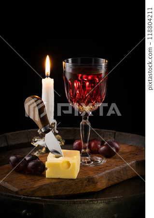 A glass of red wine with a bottle, a piece of cheese and grapes on an old wine barrel and a candle A glass of red wine with a bottle, a piece of cheese and grapes on an old wine barrel and a candle 118804211