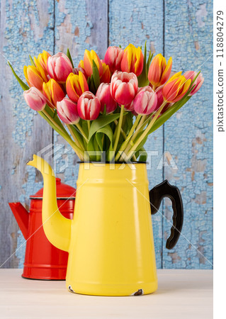 Fresh colorful bouquet of tulips in yellow enamel coffee pot in front of a blue old wooden wall and in the background still a second red coffee pot 118804279