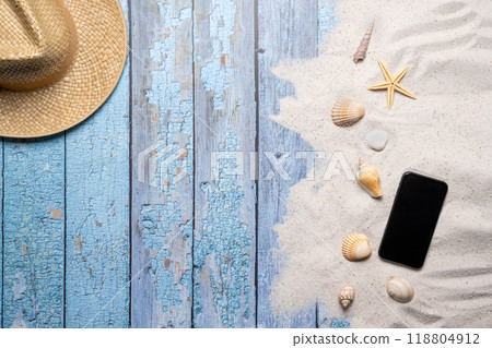 Summer, beach and vacation concept with free text space. Top view. Flat layout with a man straw hat, smartphone, various sea shells and fine beach sand on an old blue wooden boards background Summer, beach and vacation concept with free text space. Top view. Flat layout with a man straw hat, smartphone, various sea shells and fine beach sand on an old blue wooden boards background 118804912