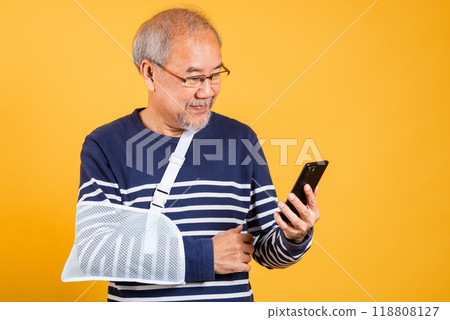 Portrait Asian old man cast his arm and holding mobile phone studio isolated yellow background, retired elderly man accident broken arm wear splint treatment, insurance medical care application Portrait Asian old man cast his arm and holding mobile phone studio isolated yellow background, retired elderly man accident broken arm wear splint treatment, insurance medical care application 118808127