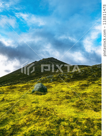 A large, rounded rock sits in the foreground of a grassy hill. The hill slopes upward towards a mountain summit under a partly cloudy sky. A large, rounded rock sits in the foreground of a grassy hill. The hill slopes upward towards a mountain summit under a partly cloudy sky. 118811478