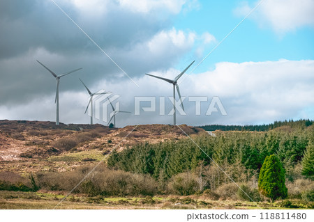 Wind turbines tower over a hilly terrain, harnessing wind energy. The backdrop features rich vegetation and a dramatic sky, showcasing sustainable energy in nature. 118811480