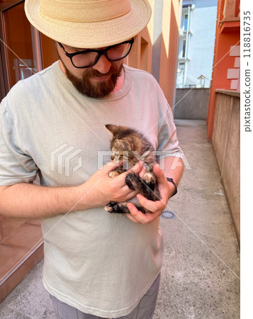 Millennial man in a straw hat and sunglasses holds lovingly at a small kitten in his hands. Tenderness moment captures the bond between human and pet. Themes of care and affection 118816735