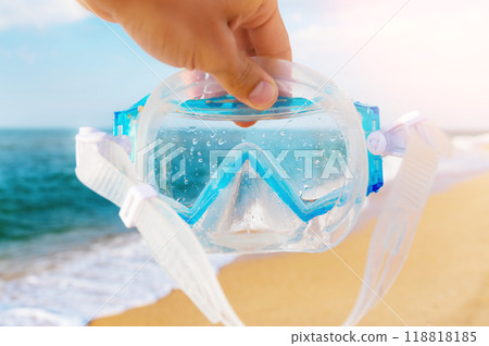 A man holds a snorkel mask in his hand against a clear blue sky and turquoise ocean. Diving mask in a man's hand. Photo of a mask and snorkel for swimming in a pool or sea with a central composition A man holds a snorkel mask in his hand against a clear blue sky and turquoise ocean. Diving mask in a man's hand. Photo of a mask and snorkel for swimming in a pool or sea with a central composition 118818185