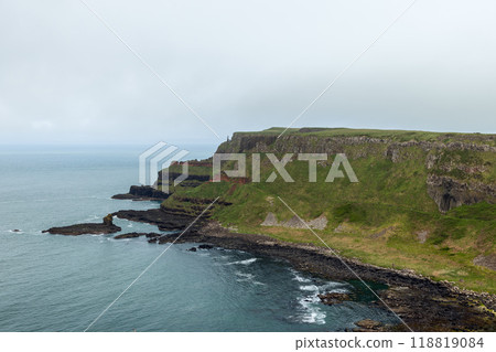 Giant Causeway cliffs with basalt rock layers and green hills meeting calm Atlantic Ocean 118819084