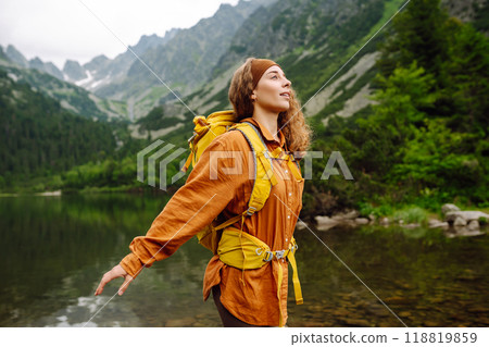 Female traveler with yellow hiking backpack against the backdrop of mountain lake with hiking poles. Female traveler with yellow hiking backpack against the backdrop of mountain lake with hiking poles. 118819859