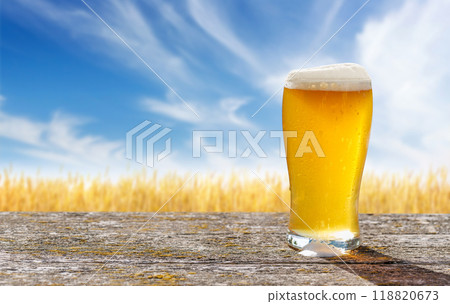 A glass of cold beer sits on a table with a vast rye field A glass of cold beer sits on a table with a vast rye field 118820673