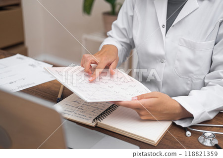 Cardiologist's Review: In a close-up view, a doctor meticulously examines papers containing patient cardiogram ECG test results, sitting at a table with a computer. Cardiologist's Review: In a close-up view, a doctor meticulously examines papers containing patient cardiogram ECG test results, sitting at a table with a computer. 118821359