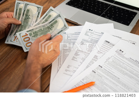 Man freelancer holds dollar bills in hands counting income for filling tax forms to report income meeting deadlines in accordance with strict laws at desk with laptop Man freelancer holds dollar bills in hands counting income for filling tax forms to report income meeting deadlines in accordance with strict laws at desk with laptop 118821403