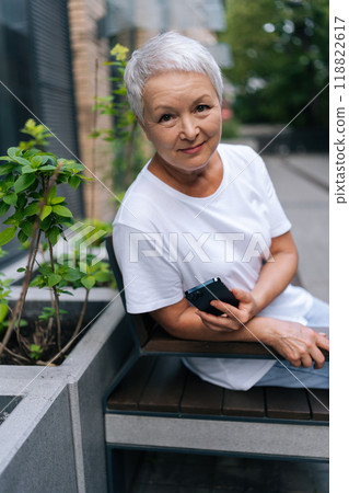 Vertical portrait of pretty senior woman wearing casual clothing holding smartphone while sitting on city bench in urban environment, smiling looking at camera with friendly expression. 118822617