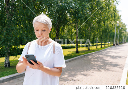 Pretty senior aged woman typing on smartphone while walking in city park. Beautiful gray-haired older woman using tapping mobile phone, smiling enjoying online communication. Pretty senior aged woman typing on smartphone while walking in city park. Beautiful gray-haired older woman using tapping mobile phone, smiling enjoying online communication. 118822618
