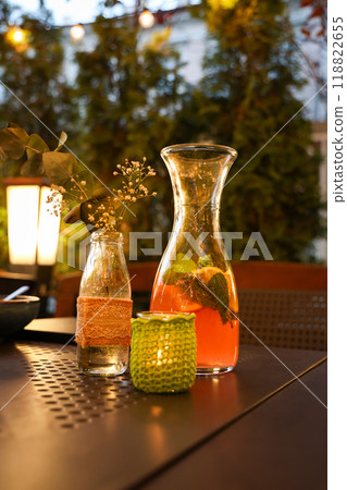 Outdoor evening scene with a glass jug of lemonade, candle in a crochet holder, and a vase with flowers. 118822655