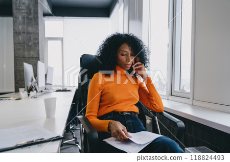 A concerned environmental analyst in a bright orange sweater delves into research reports during a crucial phone A concerned environmental analyst in a bright orange sweater delves into research reports during a crucial phone 118824493