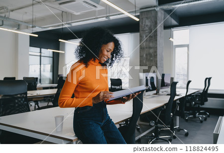 Smiling Black woman in an orange sweater reviews documents in a well-lit, modern office space 118824495