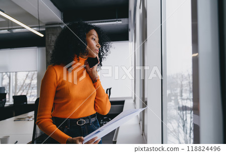 A business strategist in a vivid orange sweater ponders over a new sustainability initiative while on a call 118824496