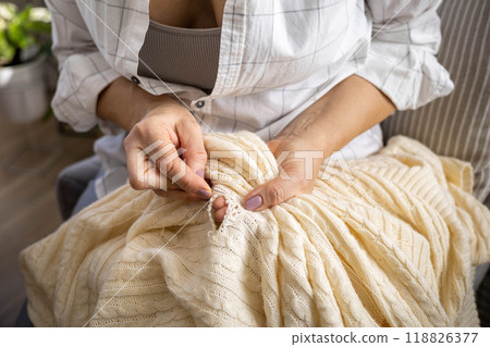 Clothes repair concept. A woman uses her hands to sew up a hole in a beige knitted garment Clothes repair concept. A woman uses her hands to sew up a hole in a beige knitted garment 118826377