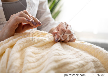 Clothes repair concept. A woman uses her hands to sew up a hole in a beige knitted garment 118826385