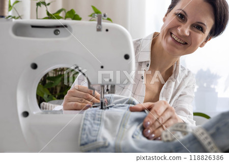 A woman at home repairs old torn jeans on a sewing machine. A woman at home repairs old torn jeans on a sewing machine. 118826386