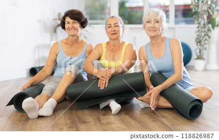 Mature women posing in seated position holding yoga rugs in fitness studio Mature women posing in seated position holding yoga rugs in fitness studio 118826987