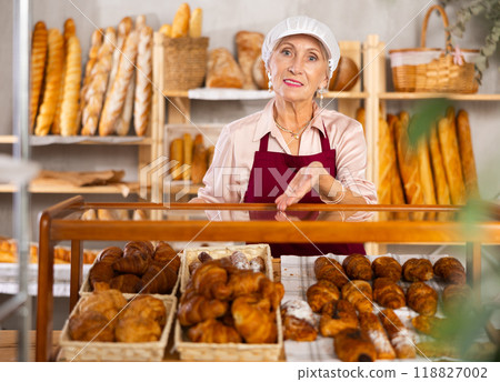 Portrait of elderly woman baker standing behind display case with fresh croissants. Offering to buy fresh baked goods 118827002