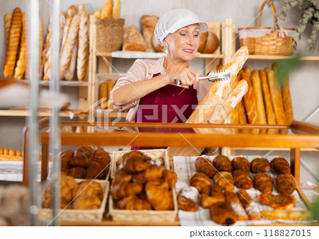Elderly woman works as salesperson in bakery and packs baguettes in paper bag for customer 118827015