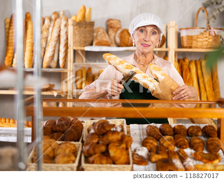 Elderly woman works as salesperson in bakery and packs baguettes in paper bag for customer 118827017