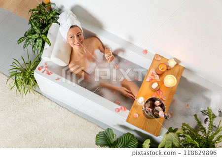 A young beautiful woman lies in a bath with foam and rose petals, reads a book and drinks champagne. A young beautiful woman lies in a bath with foam and rose petals, reads a book and drinks champagne. 118829896