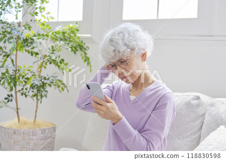 Senior woman holding her head while using a smartphone in the living room 118830598