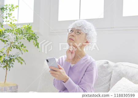 Senior woman thinking while using a smartphone in the living room Senior woman thinking while using a smartphone in the living room 118830599
