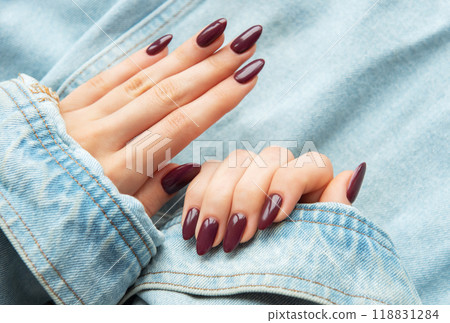 A close-up of elegantly manicured hands showcasing deep burgundy nail polish against a casual denim background 118831284