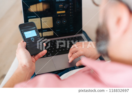 Man with notebook and smartphone sitting on sofa, online banking, internetbanking at home throught website and app. Paying with smartphone online, contactless payments by mobile phone, virtual or 118832697