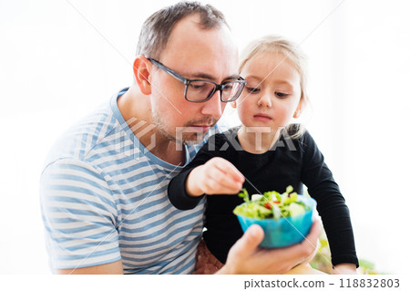 Dad and girl eating vegetables from small plastic bowl. Dad and girl eating vegetables from small plastic bowl. 118832803