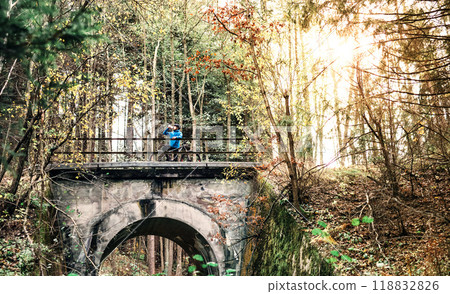 Cyclist resting on bridge and drinking water from bottle in the middle of nature, early autumn morning. Concept of healthy lifestyle. Cyclist resting on bridge and drinking water from bottle in the middle of nature, early autumn morning. Concept of healthy lifestyle. 118832826