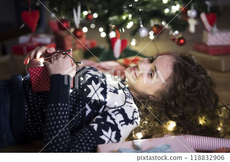 Woman lying in the middle of wrapped Christmas presents by Christmas tree. Preparing for Christmas Eve. 118832906