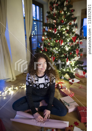 Young woman wrapping Christmas presents, sitting on the floor by Christmas tree. Preparing for Christmas Eve. 118832921